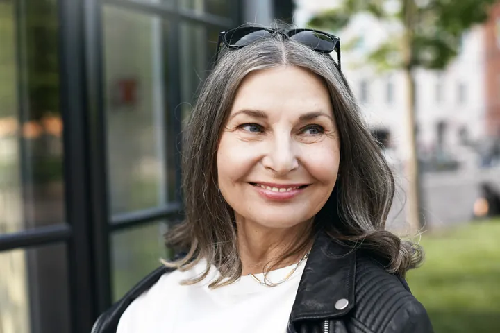 A woman in her sixties smiling warmly on a tree-lined city street
